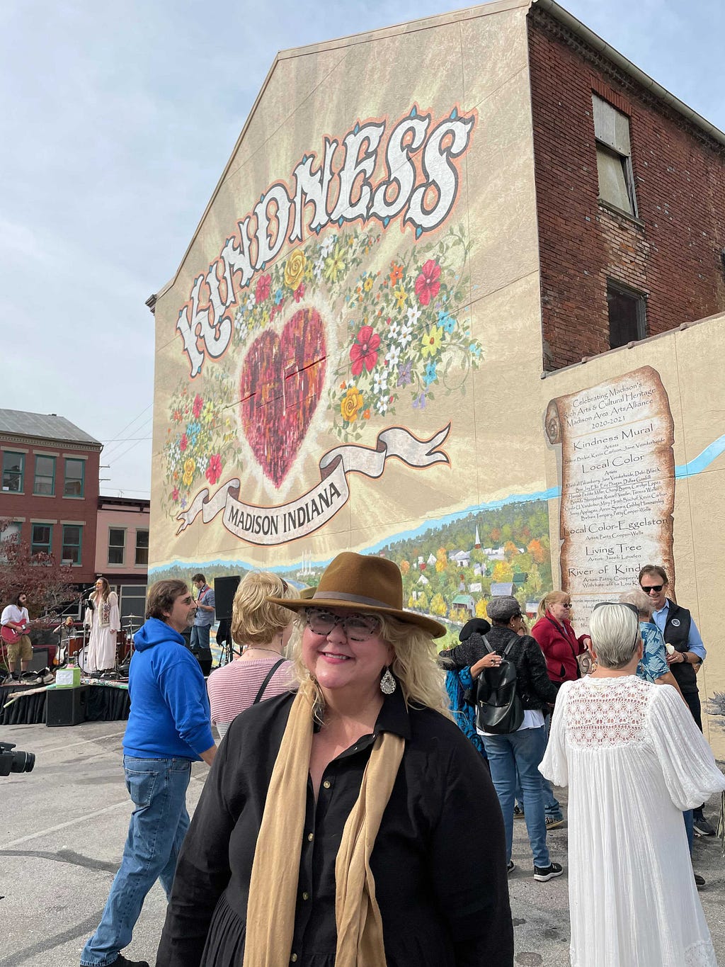 Kim Franklin Nyberg stands in front of a larger mural stating “Kindness — Madison, Indiana” surrounded by flowers and a large heart in the center.