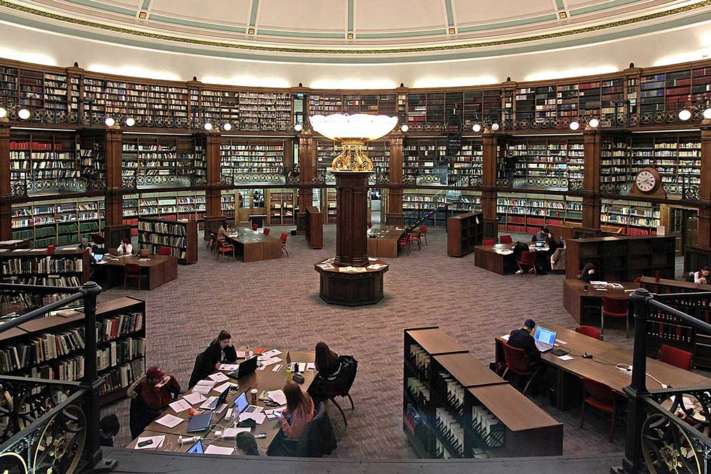 A top-down shot of the library. Bookcases positioned in an oval in the far back with many books. A large light stand in the middle of the room and large tables at the bottom with a few students studying.