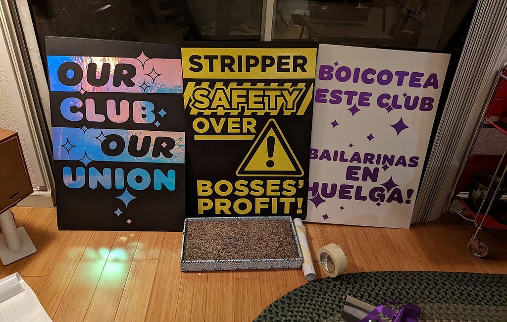 Three recently constructed picket signs sit in a domestic apartment, with craft supplies and a cat scratcher nearby. They read “OUR CLUB OUR UNION” in a holographic material, “STRIPPER SAFETY OVER BOSSES’ PROFIT!” with a caution tape theme, and “BOICOTEA ESTE CLUB BAILARINAS EN HUELGA!”, which is Spanish for “Boycot this club, dancers on STRIKE!”.