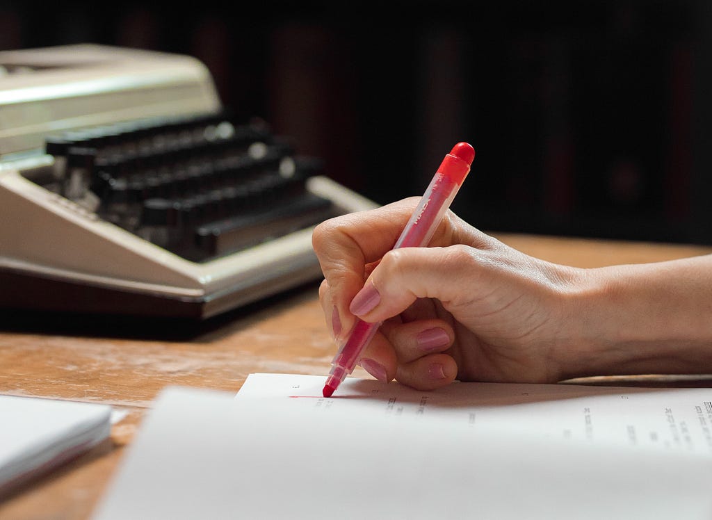 A hand holds a red pen above a manuscript, which sits on a wooden table. There is a typewriter in the background.