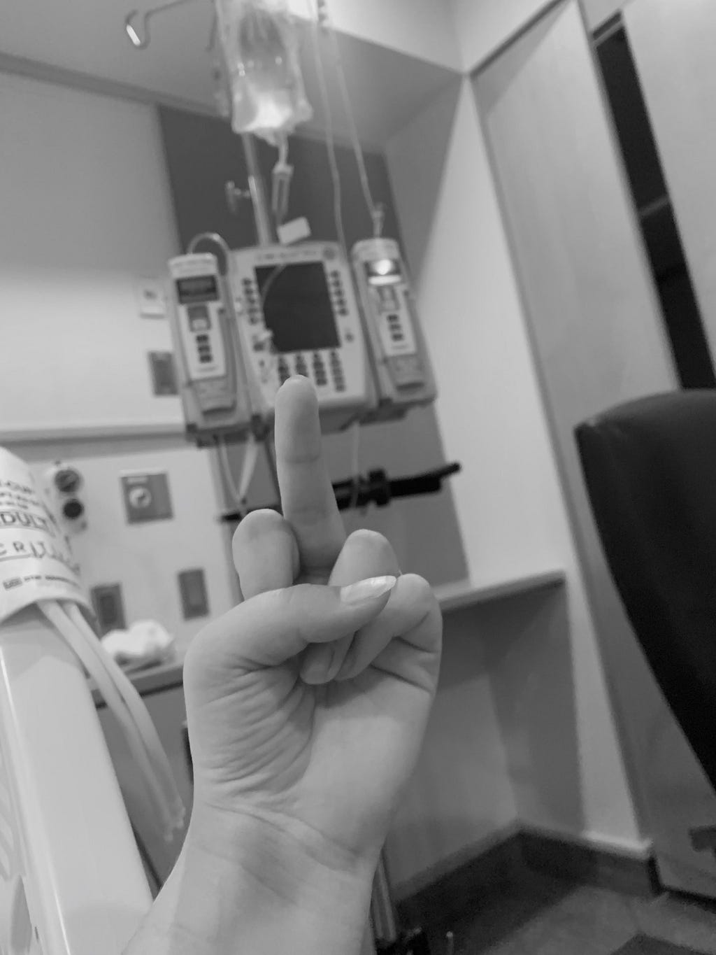 The writer’s hand is at the center focus, flipping off the IV stand behind it next to a hospital bed