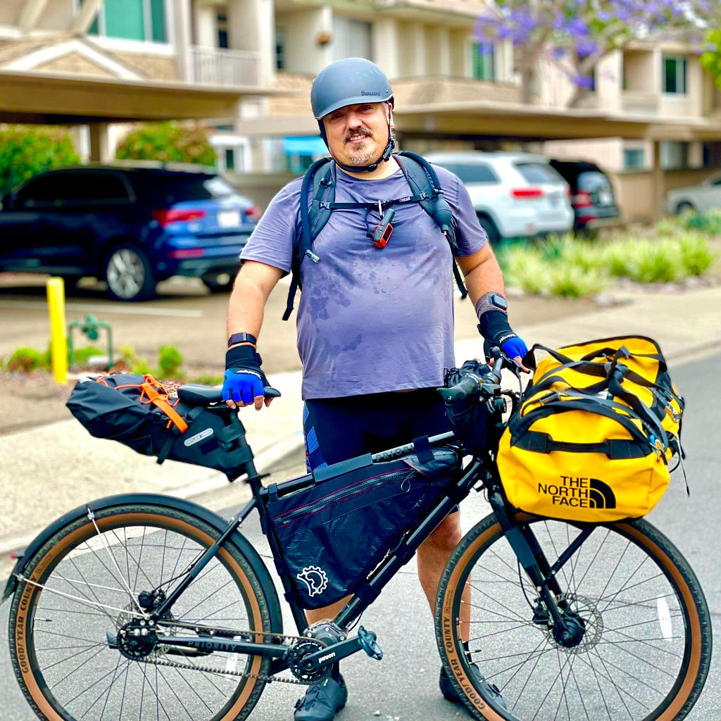 Man standing behind a bicycle loaded with bags and other gear.