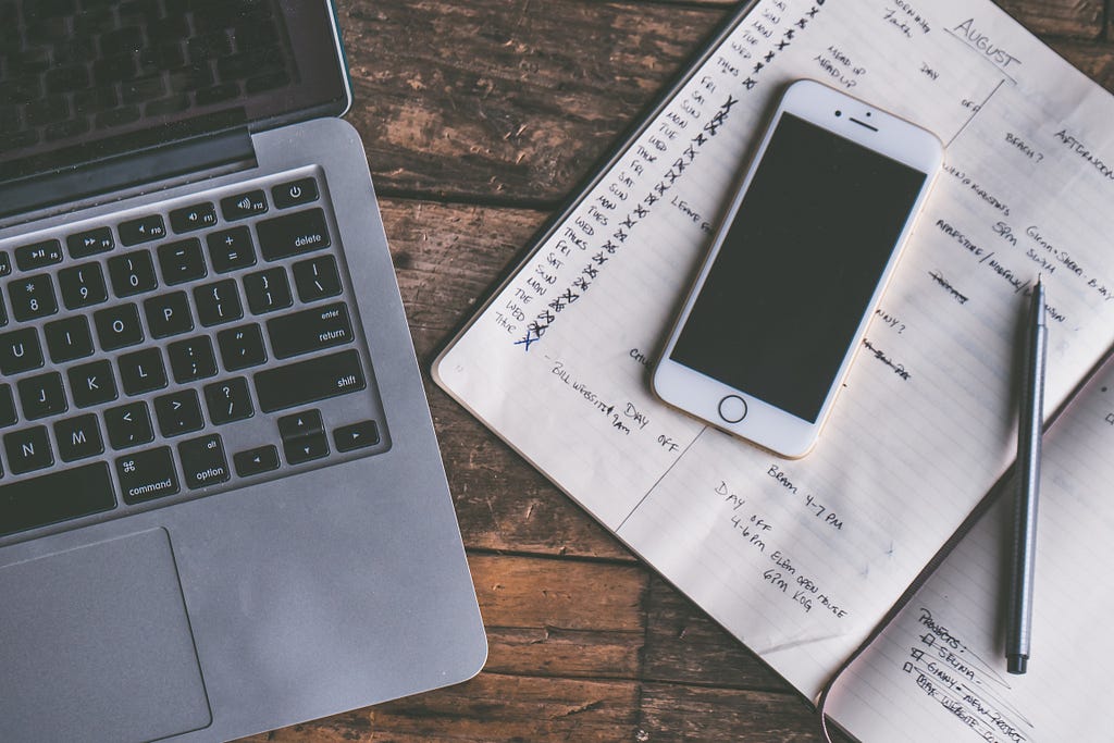 A computer, phone, pen, and planning notebook sit on a wooden table.