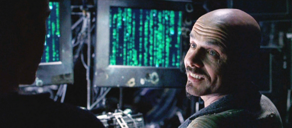 A still from the movie, Matrix, showing Cipher in front of monitors displaying arcane data dumps, and saying “You get used to it. I don’t even see the code anymore. All I see is blonde, brunette, redhead,” or something like that.