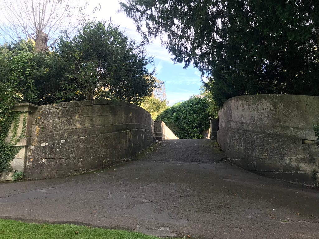 Two parapets of the railway bridge are either side of the image. They are large and very grey. They curve in from each side and funnel your view up to the narrow metal railway bridge crossing over the Great Western Railway. The path is tarmac and it takes up at least half of the picture as it is in the foreground and then narrows as it goes onto the bridge. there are trees hanging over the stone parapets either side.