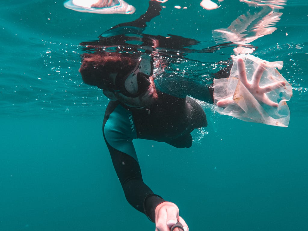 A diver in a suit and goggles, his hand reaching for plastic debris