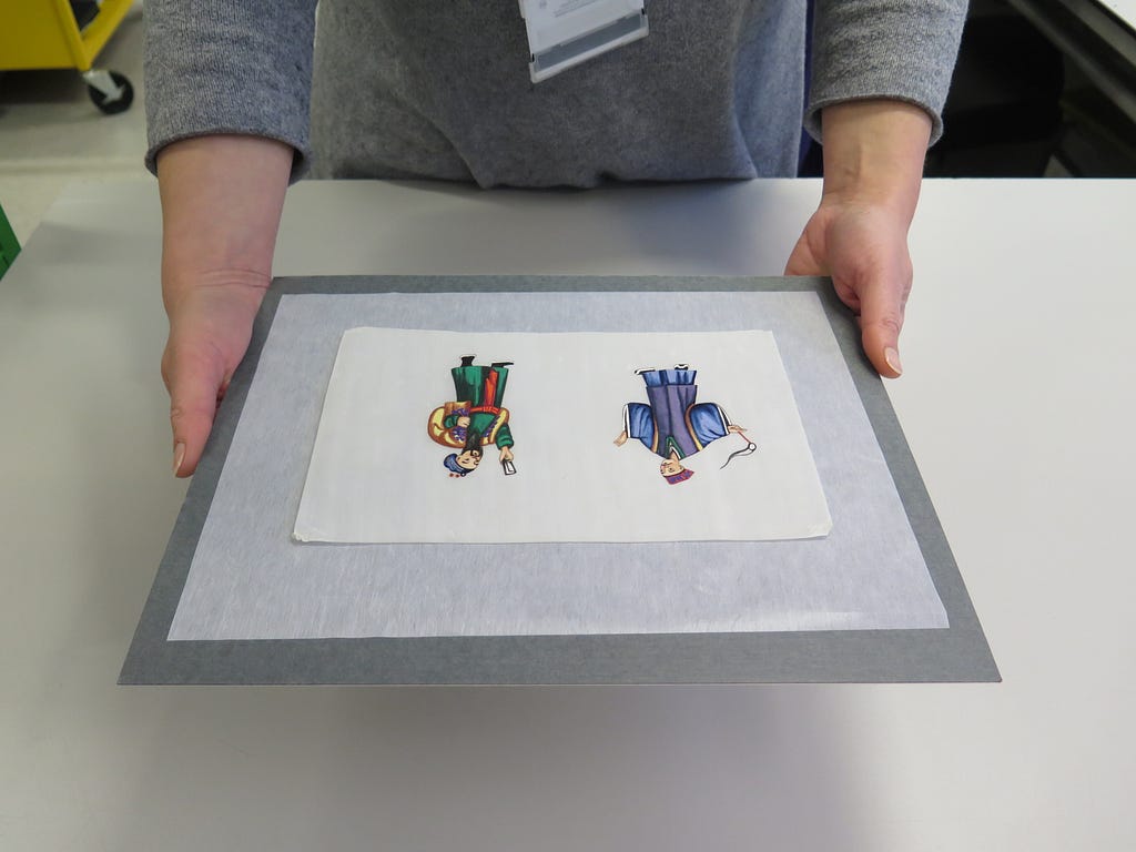 A conservator handles a Chinese pith painting by placing it on a rigid piece of archival card which she holds at the left and right edges.