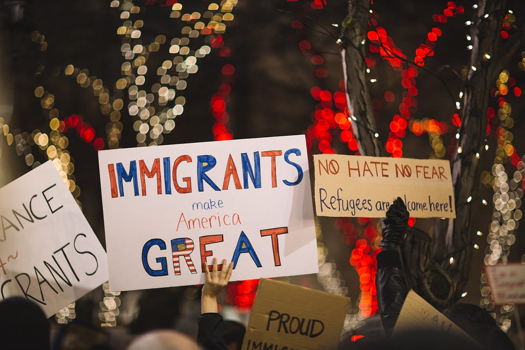 Protest signs at night. The signs read "imigrants make America great", "no hate no fear, refugees are welcome here" and "proud".