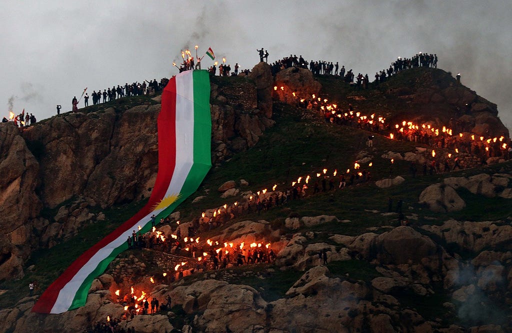 A crowd of people dotted along a large rock formation and zigzagging down in lines that are fringed by embers of flames (being held up on sticks by Kurdish people). Trailing down the mountain and held at the top is a long strip of cloth representing the flag of Kurdistan.