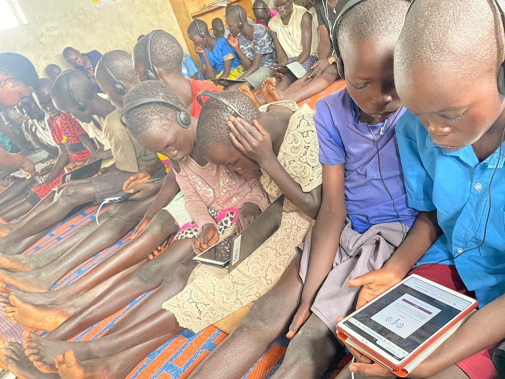 Children are sitting on the floor with their legs extended, side by side on a long row. They have headphones on and they’re holding a tablet, where they engage with Kolibri, Learning Equality’s edtech tool for teaching and learning with tech but without the Internet.