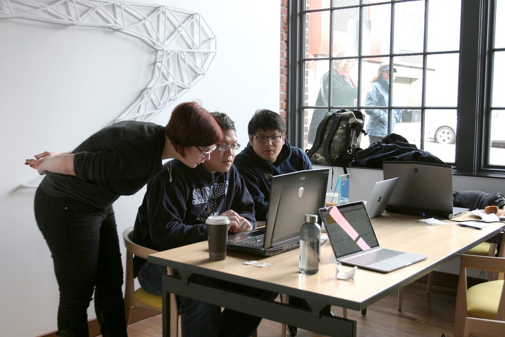 Woman with short red hair leaning over helping two men with laptops.