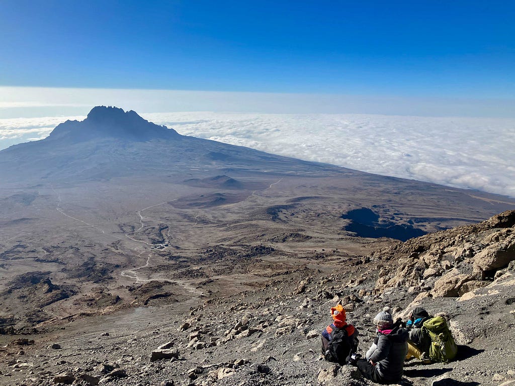 Climbers sitting on the ground on a slope of gravel with a long-distance view of terrain and clouds below, with deep blue sky on the horizon.