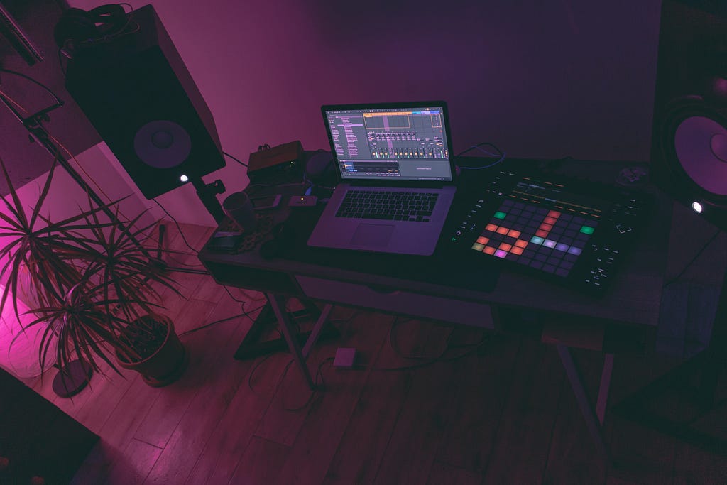 Foto de uma mesa com equipamentos de produção musical e um notebook. Ao redor, na sala, estão caixas de som e uma planta.