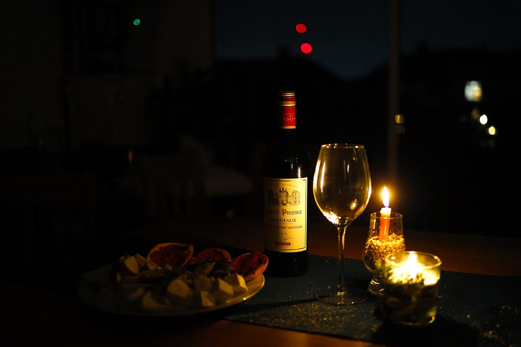 A bottle of wine, lit candle, empty wine glass and some food on a table.