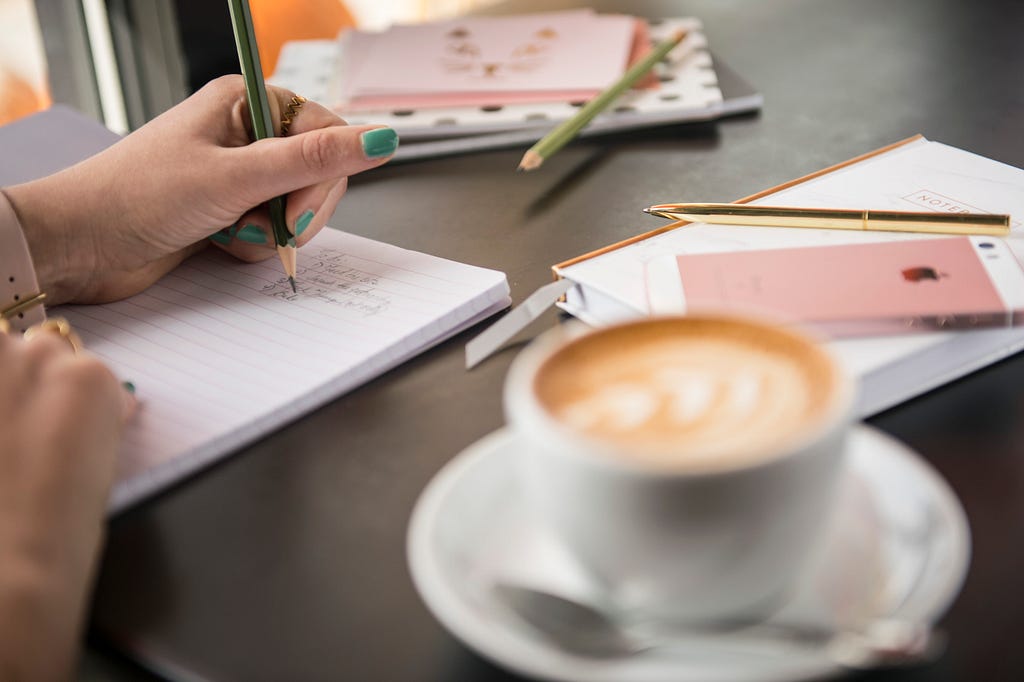 A hand writing in a notebook, with a latte in the foreground.