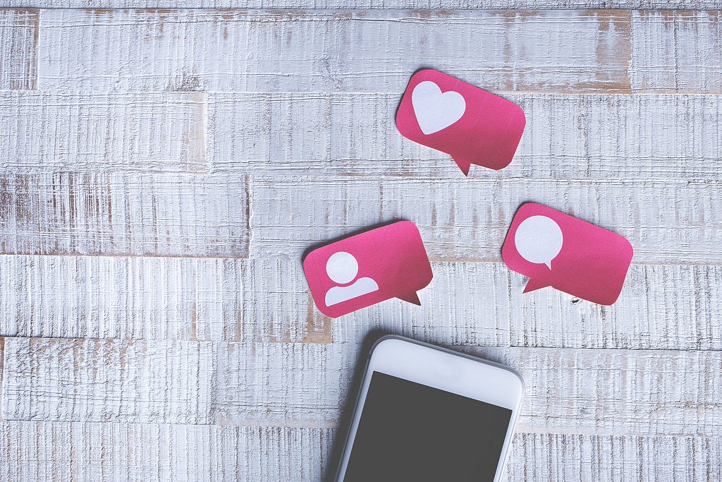 White smartphone on a white wooden table surrounded by three pink paper icons shaped as speech balloons.