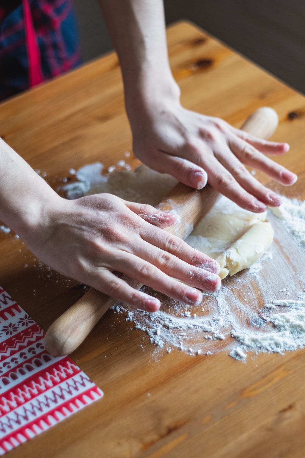 Two hands rolling out dough with a wooden rolling pin on a wooden table covered in flour.