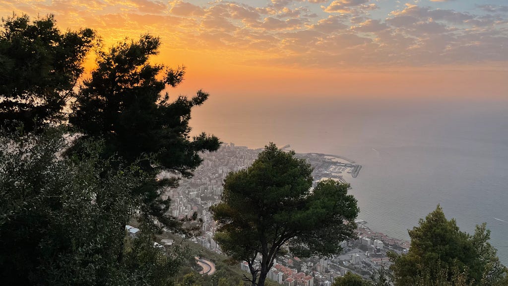 A picture overseeing Beirut from afar. Trees taking place at the forefront, the city is grey just like the sea, the sky vibrant orange with a few clouds.