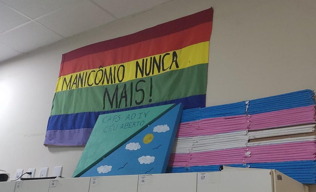 Bandeira arco-íris com os dizeres “manicômio nunca mais!” escritos em tinta preta. A bandeira está pendurada em uma parede branca acima de um quadro que contém um céu azul pintado e os dizeres “CAPS AD IV Céu Aberto” escritos em tinta azul e de uma bandeira do orgulho trans feita com canudos pintados de azul, rosa e branco colados em um pedaço de papelão.