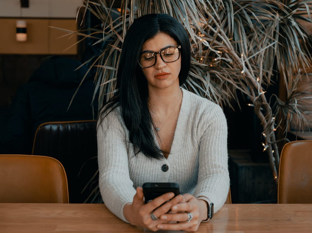 A woman with dark hair and glasses sits in an office boardroom and is distracted by her phone.