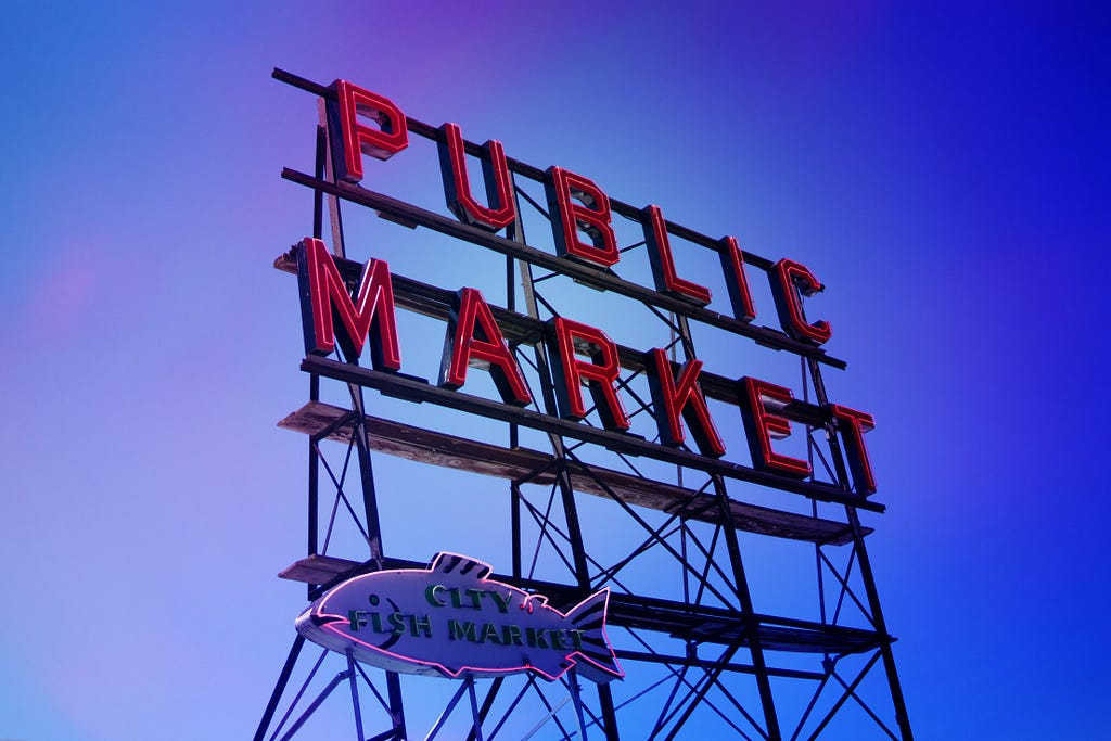 A neon signage on metal billboard stilts, with the words ‘Public Market’ can be seen. While a smaller sign designed in a shape of a fish that says ‘City Fish Market’ is placed below. The sky is a dark shade of blue, perhaps it is sundown or sunset.