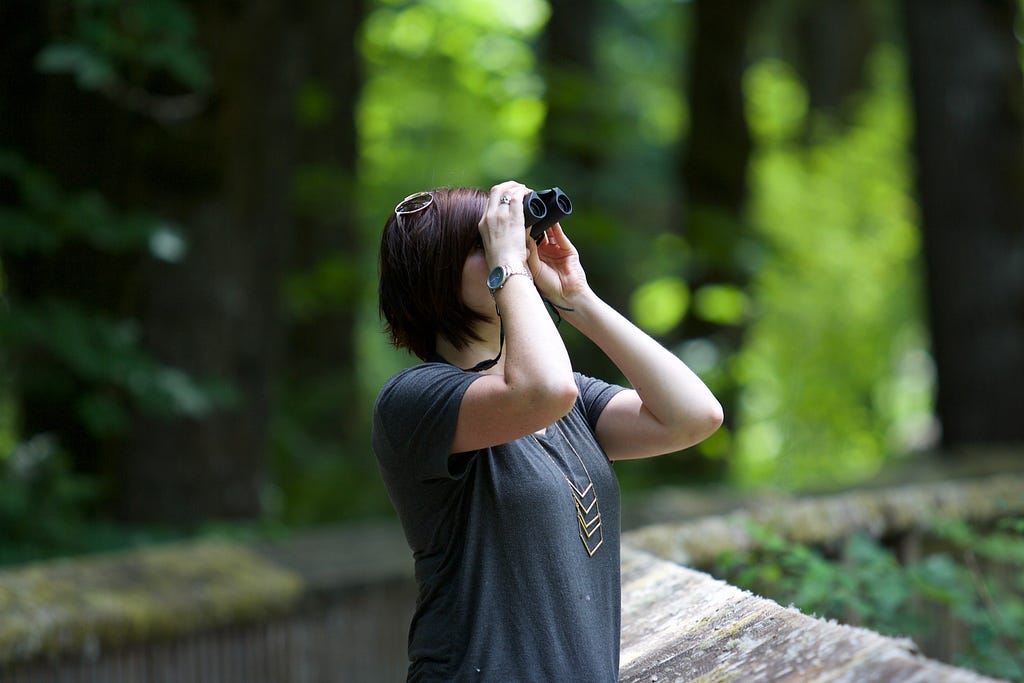 Woman in a forest looking up at trees with binoculars.