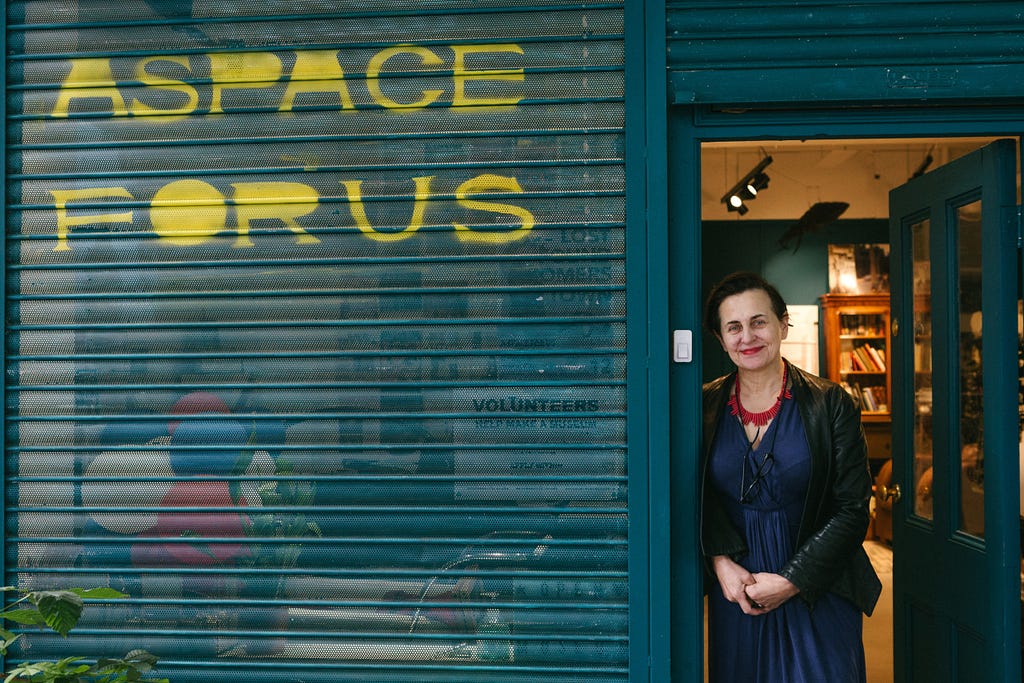 Photo of a woman standing in the doorway of a museum. On the blue museum shutters is graffiti in yellow paint: “A space for us”. The woman is white, with dark brown hair and red lipstick, wearing a blue dress, black jacket and red beaded necklace. Behind her, in the museum, we can see a bookcase.