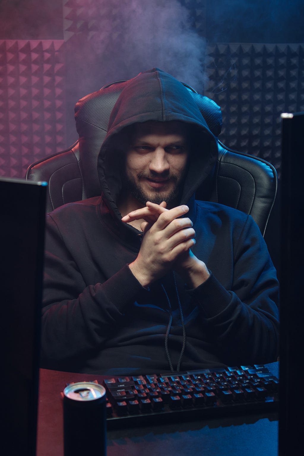 A young man in a hooded sweatshirt sits at a desk in a high-back swivel chair facing the viewer, and a computer off to one side. The room is poorly lit with eerie light, and the young man looks like he’s scheming some dastardly deed.