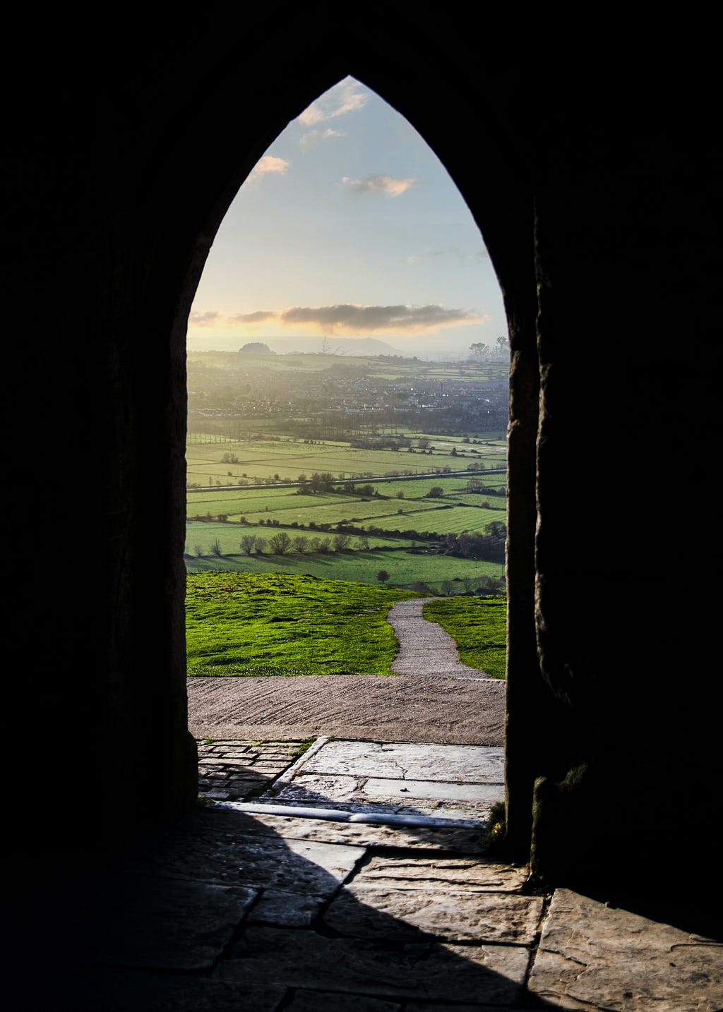 A glorious view of green fields and blue sky with white clouds can be seen through an old, church-style stone window frame.