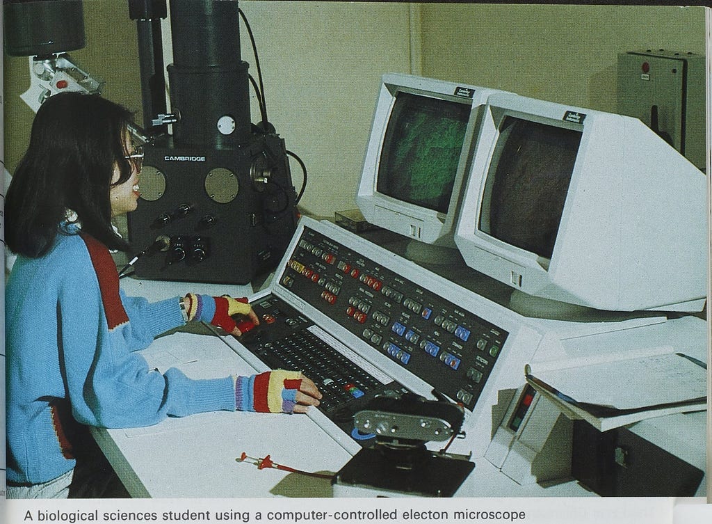 A student in striped fingerless gloves, a blue jumper and glasses sits at a desk at the keyboard of a large electronic machine with several rows of coloured buttons and two white monitors.