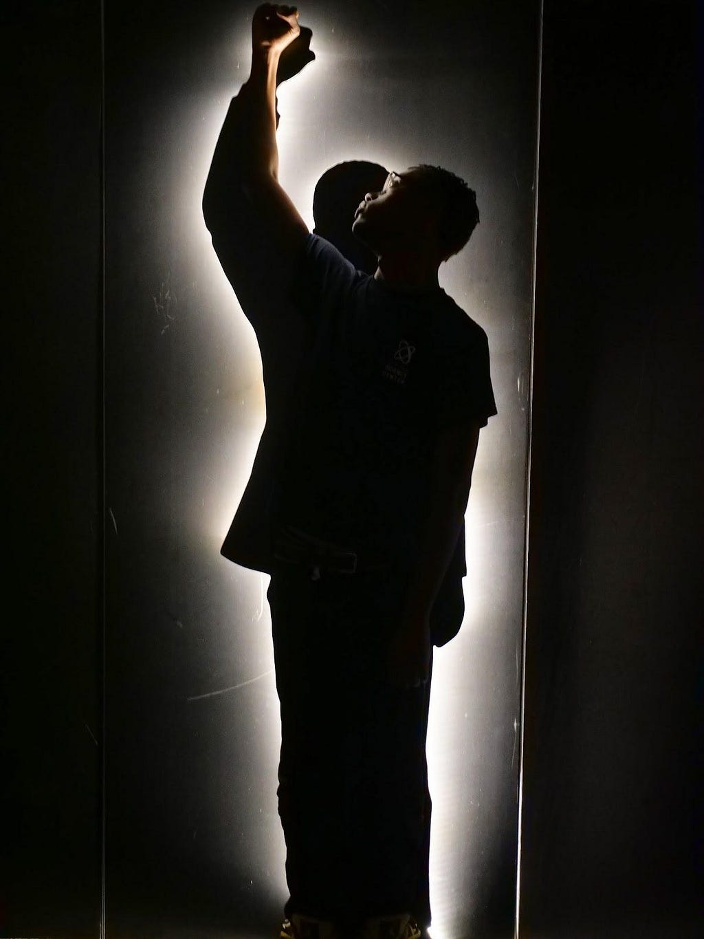 A photograph of a Black teen standing in front of a silhouette of a slightly taller Black man. Both the teen and the silhouette hold one raised fist in the air. The silhouette is outlined by white light. The teen’s back is to the viewer and he looks to the left, towards his raised fist. It is too dark to see what the teen is wearing. He wears glasses.