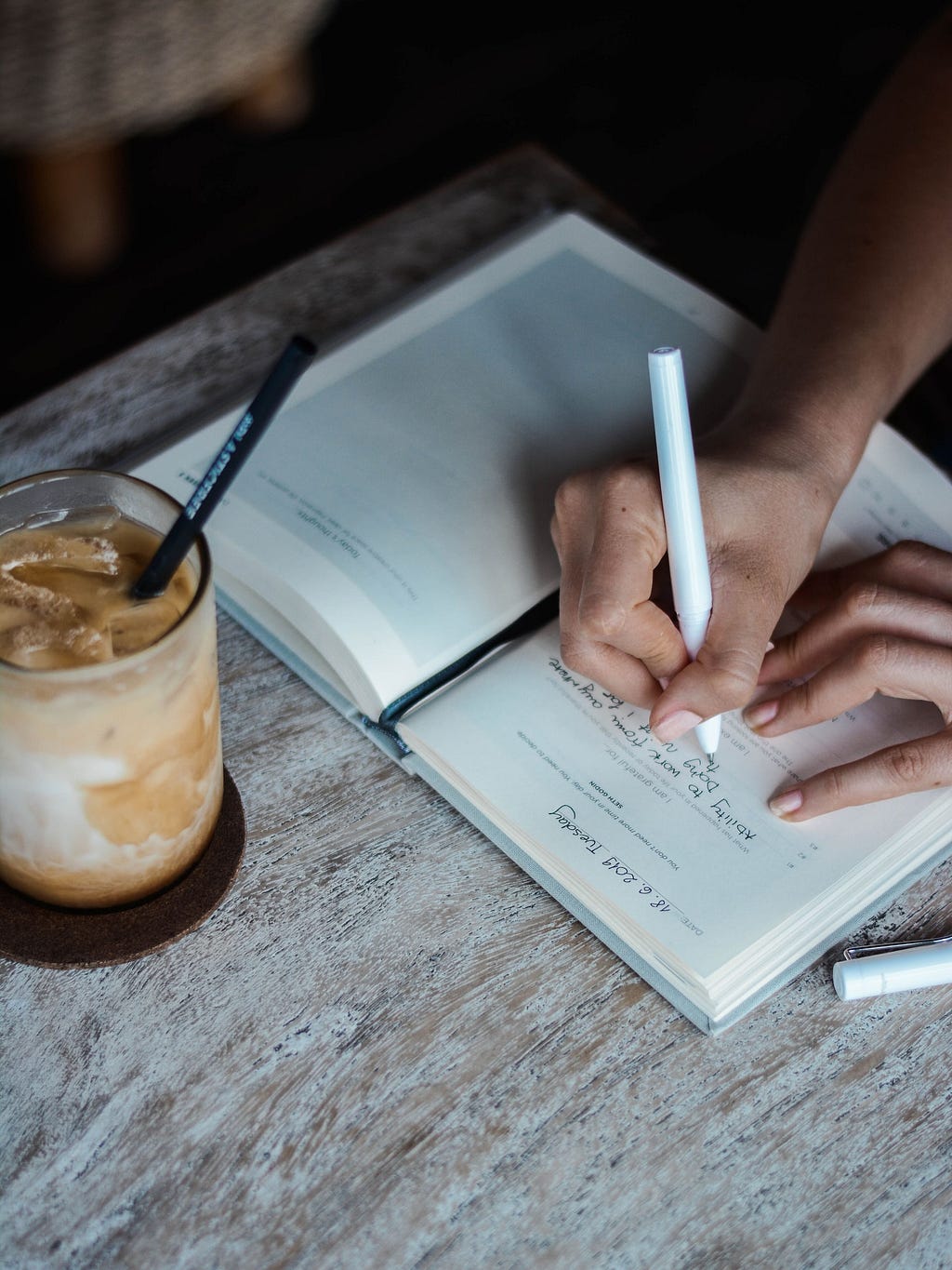 Overhead view of a woman’s hands writing in a journal with a pen. A creamy, iced drink with a straw is just above the journal.