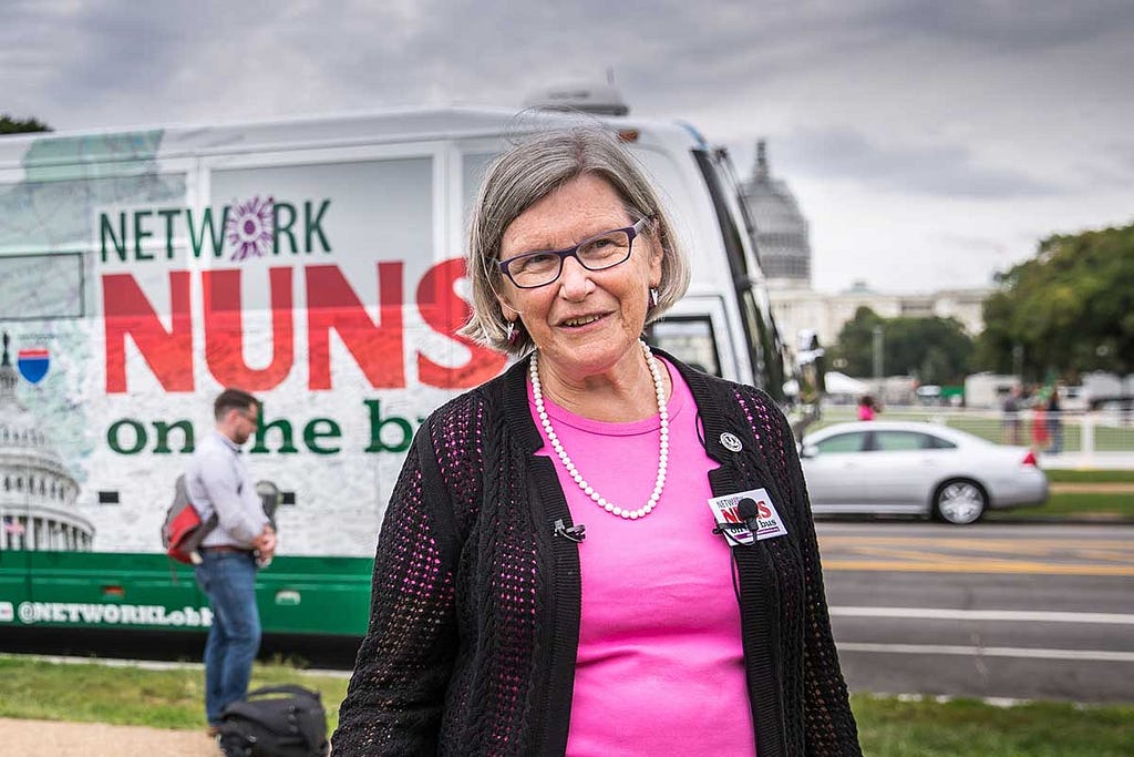 Sister Simone Campbell in pink top and black sweater stands in front of the bus bearing the logo “Nuns on the Bus.”