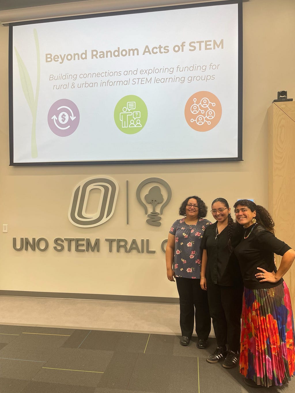 Dr. Lisette Torres-Gerald with two other people in front a sign that says Uno STEM Trail Center and their logo. There is also a projector behind them has the Beyond Random Acts of STEM logo. Dr. Torres-Gerald (Right) is wearing a gray shirt with pink flowers and black pants, Valeria Alderete (Middle) is wearing an all black button up and black pants, and Ivel Gontan (Right) is wearing a black top with a colorful floral pleated skirt.