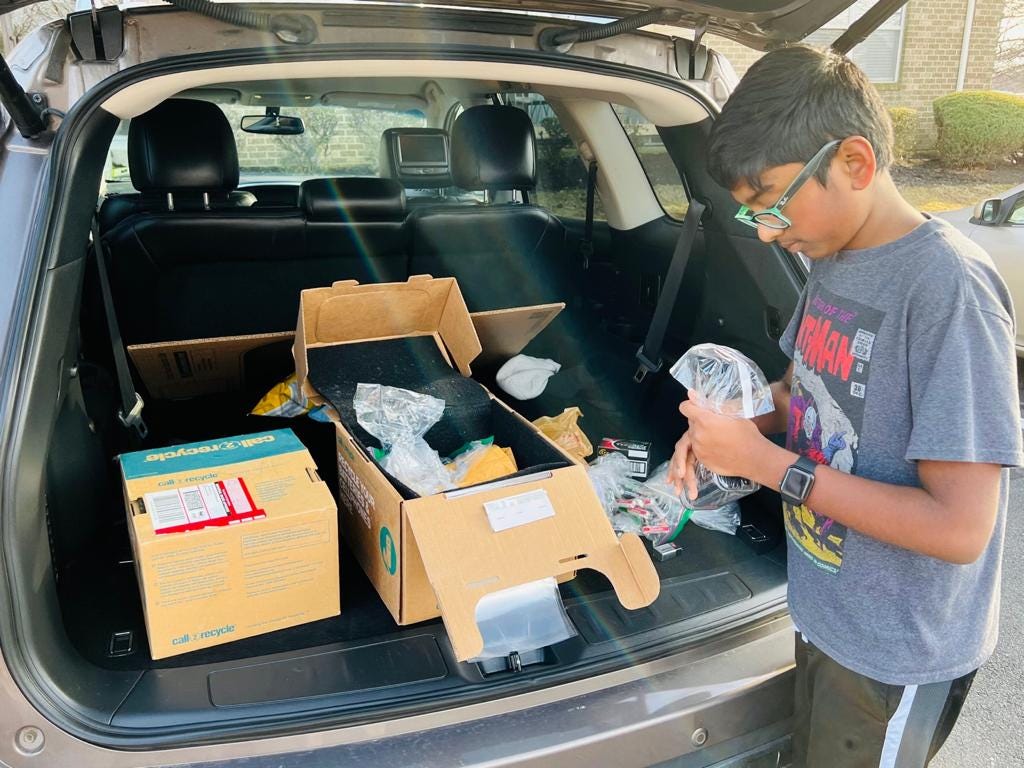 Brown boy with short black hair wearing turquoise glasses filling boxes in a trunk on a sunny day