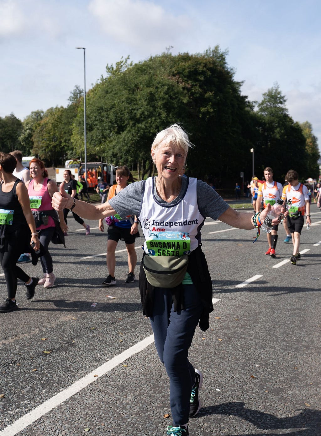 Susanna, an older woman, smiles at the camera as she runs. She wears an Independent Age running vest and holds her hands in two thumbs up.