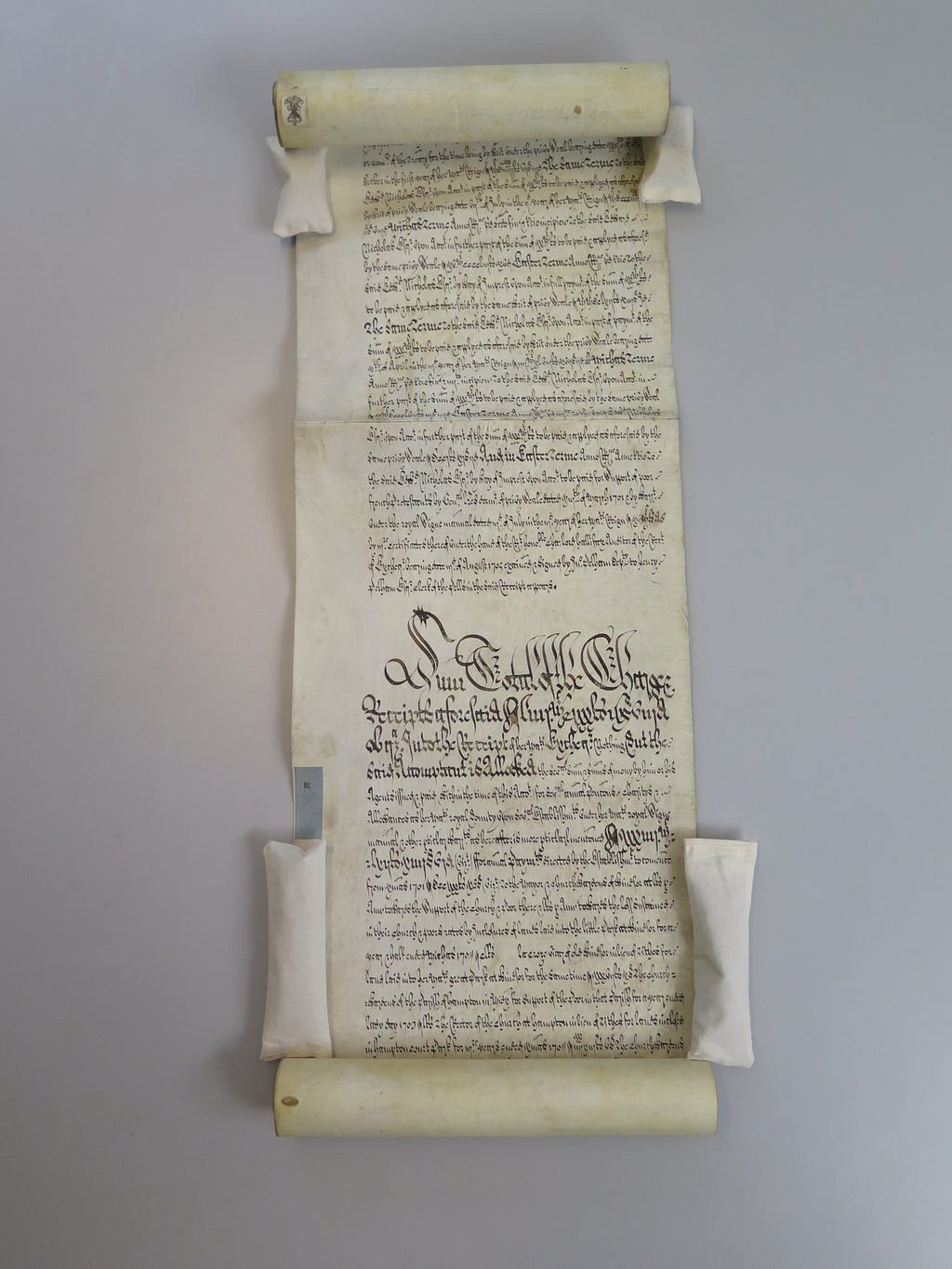 A handwritten parchment scroll which has been unfurled in the middle section and held down by soft weights along the edges.