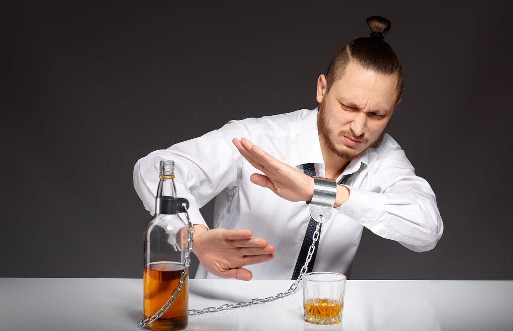 A man in a dress shirt and time sitting at a dinner table shelters himself from the glass and bottle of scotch in front of him that is shackled to his wrist