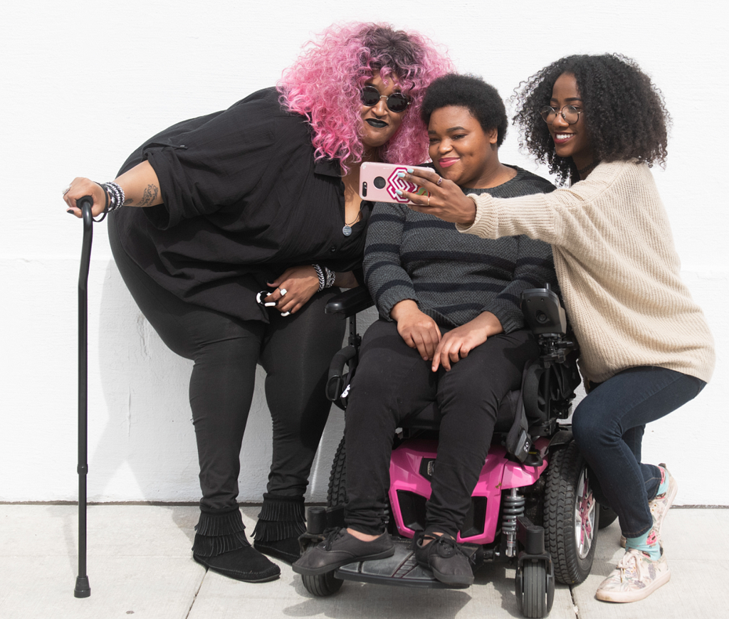 3 women leaning in together for a selfie. 1 holding a cane, 1 seated on a power wheelchair, and 1 woman hold a phone.
