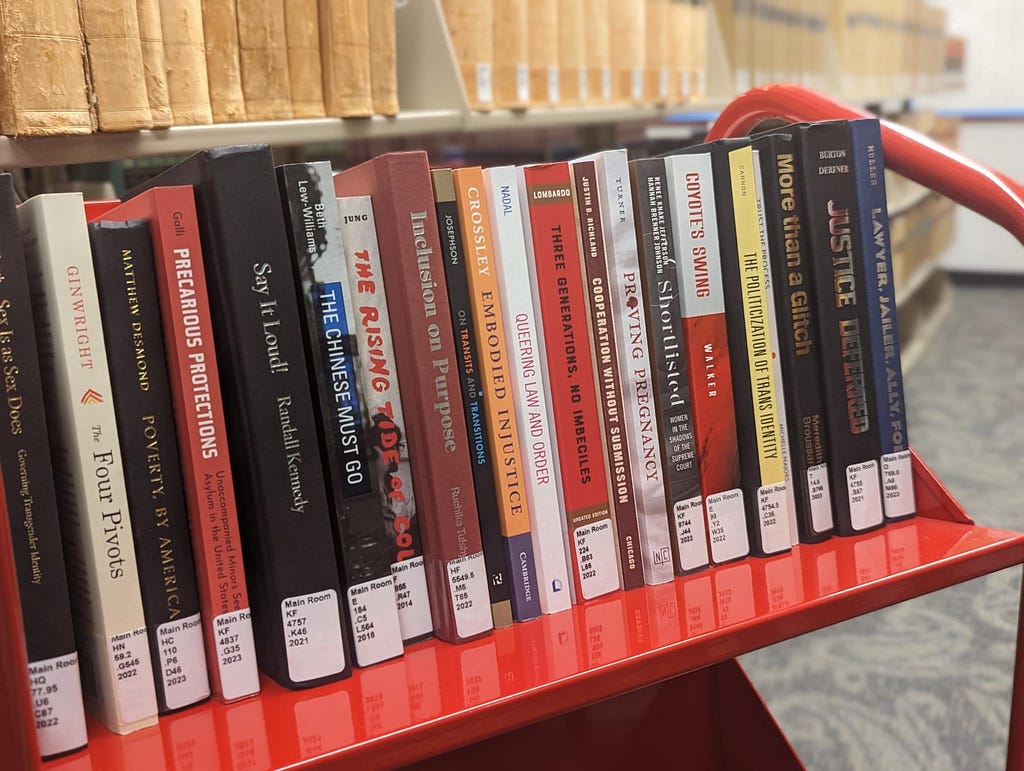 Twenty one books are lined up on the top shelf of a red book cart with their spines facing out. The book cart is placed at a slight angle in front of library shelving.