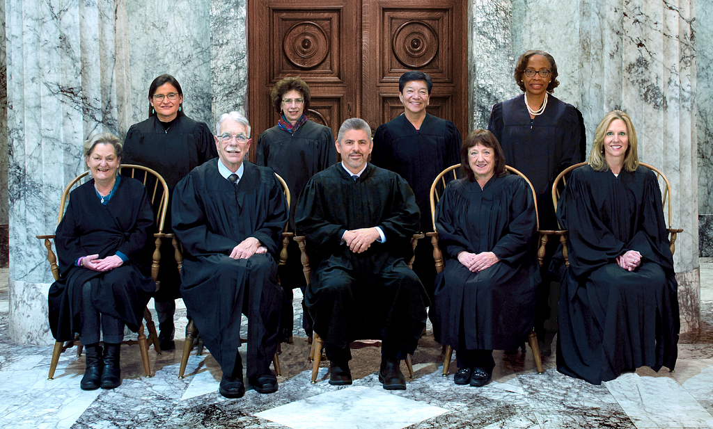 Five justices of the Washington State Supreme Court sit side by side on chairs while four more stand behind them. They are wearing their black robes and are positioned in front of the ornate wooden doors of the court room at the Temple of Justice.