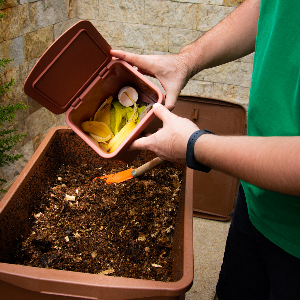 A person wearing a green shirt and a black wristband is holding a small brown compost bin filled with food scraps, including banana peels and eggshells, over a larger compost bin. The larger bin contains decomposing organic matter and has a small orange trowel resting inside it. The background features a stone wall and some greenery.