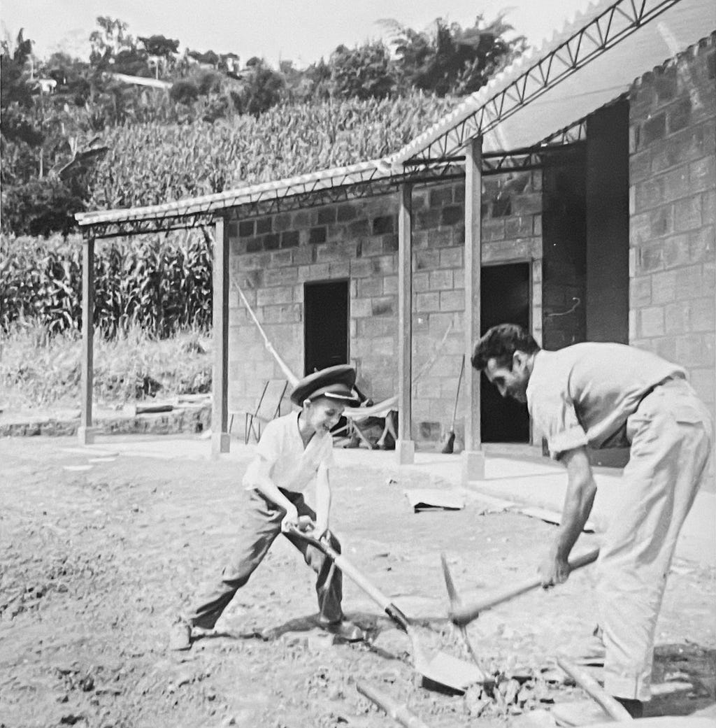 Black and white photo showing a bungalow under construction in the background; in the foreground, a child and adult playfully working the grounds with a shovel and pitchfork. Child is clearly having fun.
