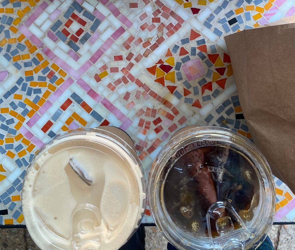 A photo of two coffees from Wine & Butter placed on a colorful mosaic table with a brown bag (with croissants inside) cut off on the right side.