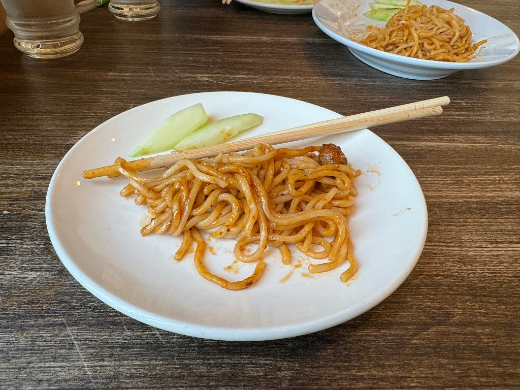A white plate of peanutty dan dan noodles and a pair of chopsticks.
