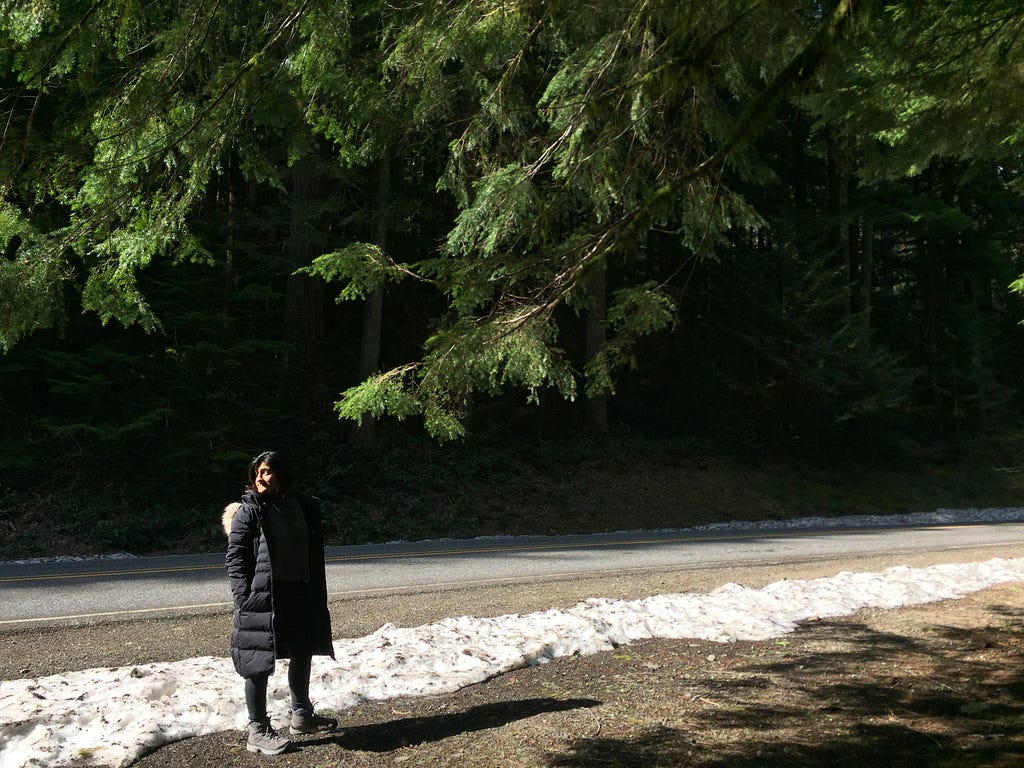 A woman stands on the side of the road with snow nearby, with the branches of an evergreen tree above her.