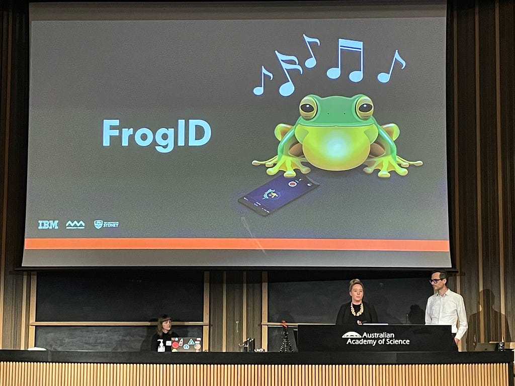 Photo showing a projection screen with a slide that says “FrogID” and shows an illustration of a frog with notes to indicate that the frog is calling and a smartphone app in front of the frog. The photo also shows Isabella Bain and Martin Tomitsch standing at the lectern in a conference  theatre space.