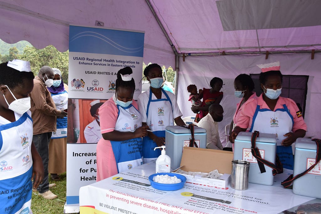 Health workers in nurses caps and masks stand under a tent covering a table filled with coolers and supplies to administer vaccinations.