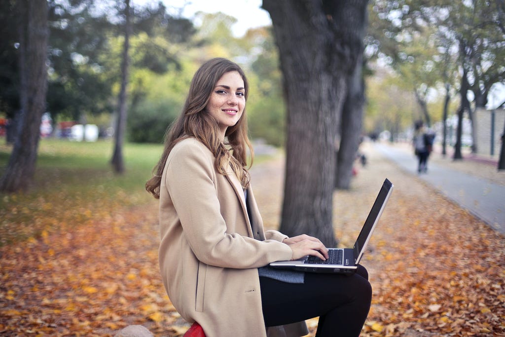 Mulher sorri, enquanto trabalha em um notebook, sentada próximo às árvores de um parque.