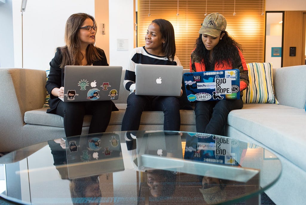 Three women of colour sat on a sofa with their laptops open with a low table in front of them